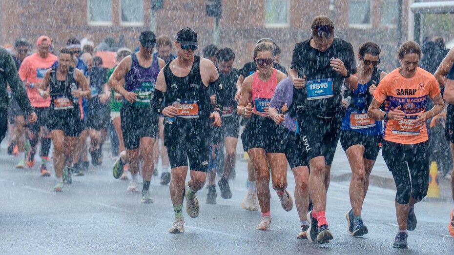 Marathon runners in Edinburgh fight through the heavy rain. They are dressed in running gear and travelling along a flat road.