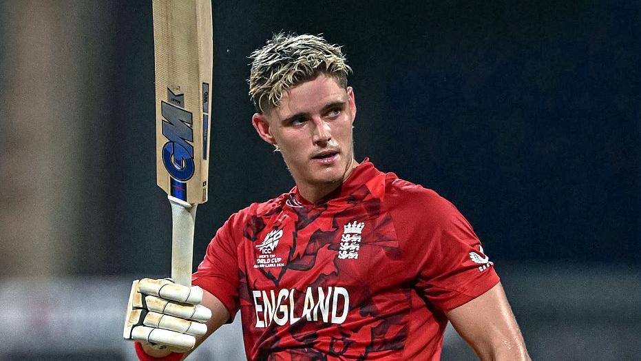 Jacob Bethell, in red team shirt, raises his bat after scoring a century for England