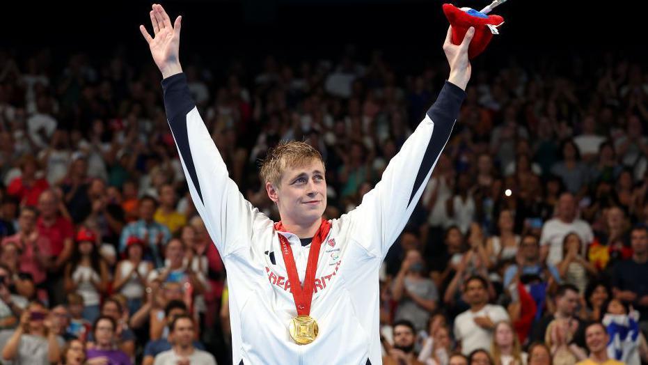 Gold medallist Stephen Clegg celebrates on the podium at  La Defense Arena, wearing his gold medal and his Team GB tracksuit, he raises his arms in the air and looks at the crowd - a sea of faces is behind him as he enjoys his moment in the arena.