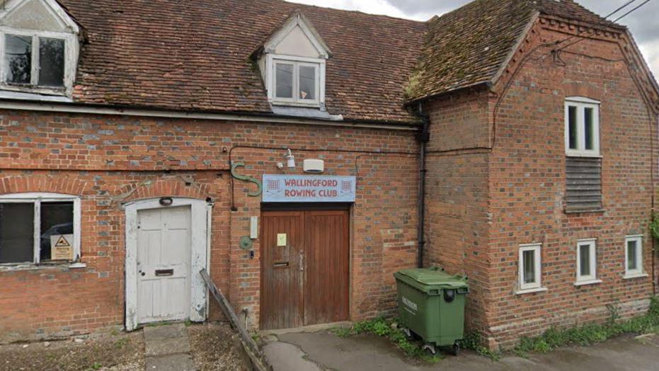 The front entrance of an old brick two-storey building. Above double wooden doors is a sign which reads: "Wallingford Rowing Club". In front of the building is a narrow pavement in front of a road with double yellow lines.