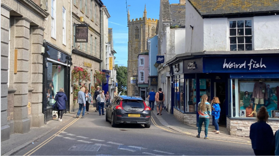 St Ives town centre with a car driving through a narrow road surrounded by people and the church in the distance 