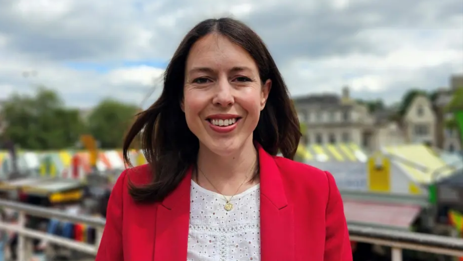 Profile picture of Alice MacDonald pictured outside Norwich Market, smiling at the camera. She has long brunette hair, red lipstick, a gold necklace, and a white top with a red suit jacket over the top. 