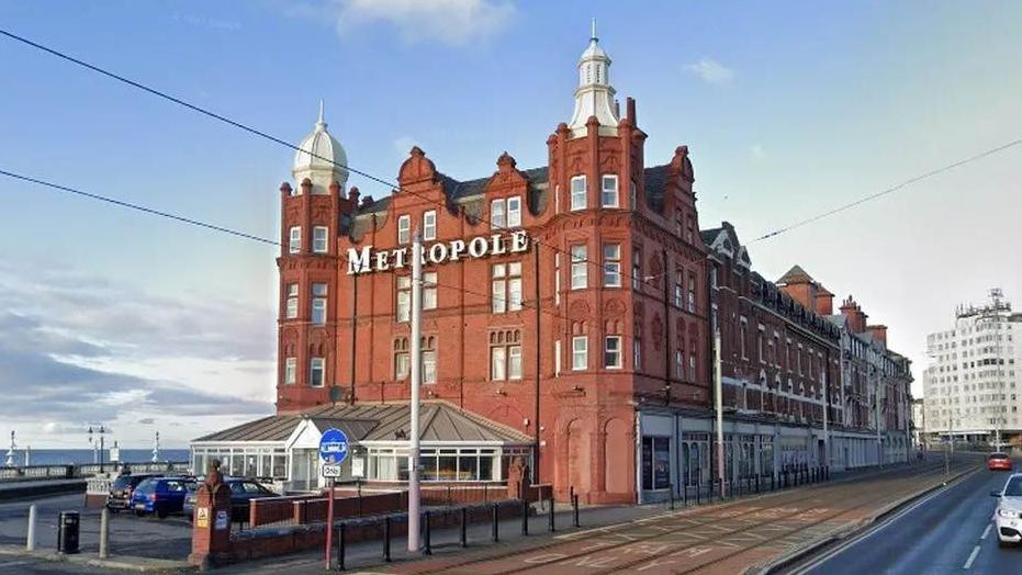 Street view image of the Metropole hotel, a large red brick building, located between tram tracks and the seafront, in Blackpool.