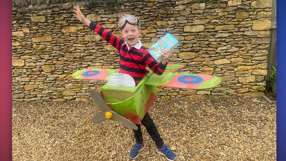 Boy in black and red striped long-sleeved t-shirt with goggles on his head and a cardboard spitfire-style homemade plane around his middle. He is holding a book of Grandpa's Great Escape by David Walliams.