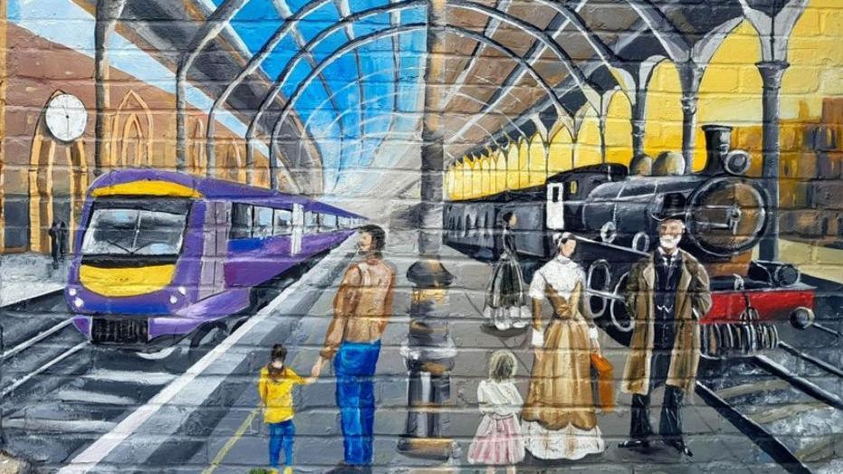 People standing on a railway platform - some in modern clothing and others in Victorian dress. There is a steam locomotive to the right of the image and a modern train to the left.