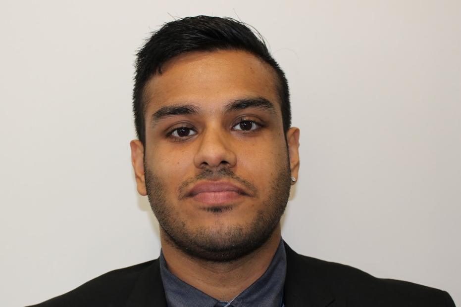 A young man with short dark hair looks at the camera with a slight smile. he stands in front of a white background and wears a black suit jacket with a dark grey shirt.