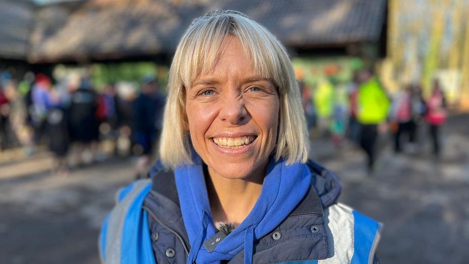 Emma Sperring looks into the camera, smiling. She is wearing a blue hoodie underneath a blue jacket and blue high-vis vest. She has a blonde bob with a parted fringe. 