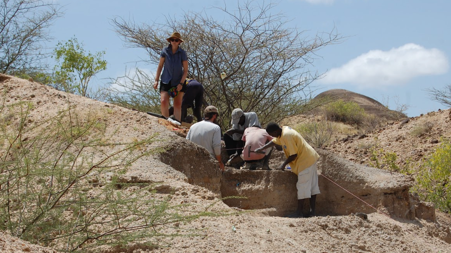 A group of people are working together at an archaeological dig site in a dry, rocky landscape with sparse vegetation. Some are kneeling and carefully examining or excavating a partially exposed layer of earth, while one stands nearby watching. Their focused activity suggests they are searching for or studying artifacts or fossils in the ground.