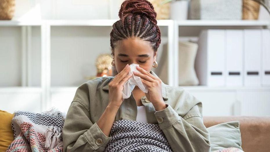 A young woman sits on her sofa, blowing her nose into a tissue