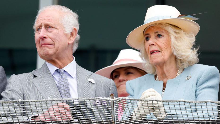King Charles III dressed in a light grey suit and tie grimaces as he sits beside Queen Camilla who wears a light-coloured fedora hat with a blue feather and a pale blue two-piece skirt suit