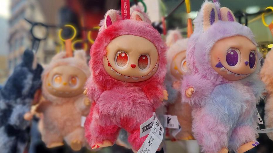 Three small furry dolls of various colours on display for sale. They have large eyes and are creature-like in appearance.