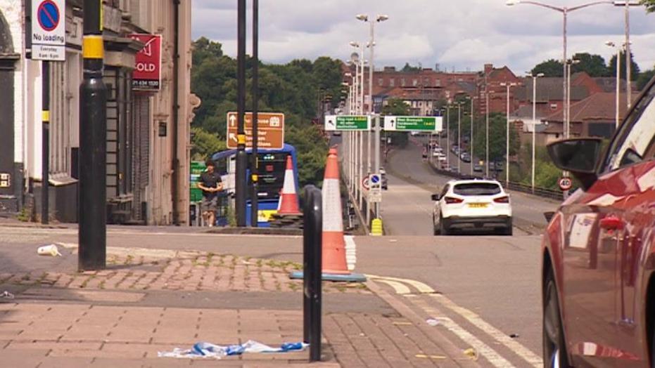 Scene of crash shows top of a slight hill with cars on the road and a police sign on the ground. 