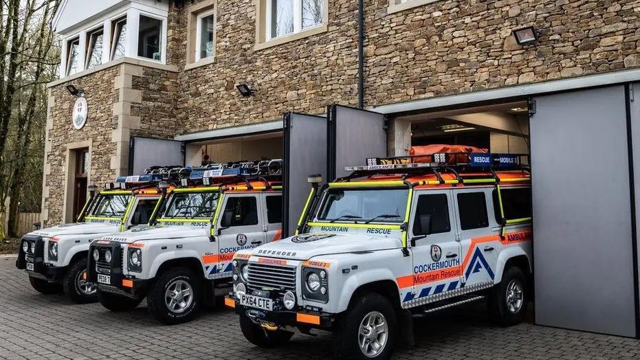 Three Land Rover Defender vehicles are parked outside Cockermouth Mountain Rescue Team's headquarters. The vehicles have emergency lettering and fluorescent strips with technical equipment attached to their roofs.