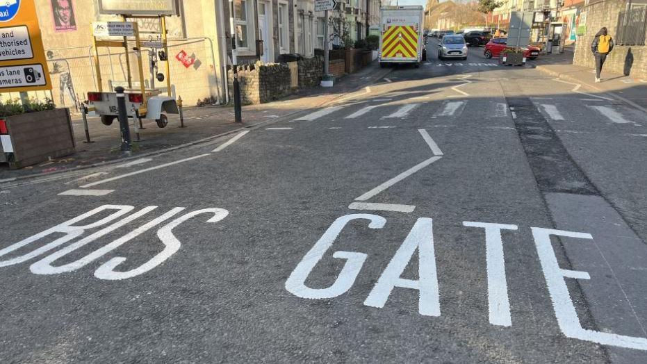 The picture shows the words "bus gate" painted on the road in white on the grey tarmac. There are buildings and vehicles in the background.