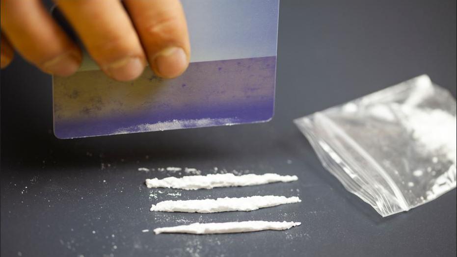 A hand holds a plastic credit card above three lines of white powder on a table top next to a small empty plastic bag