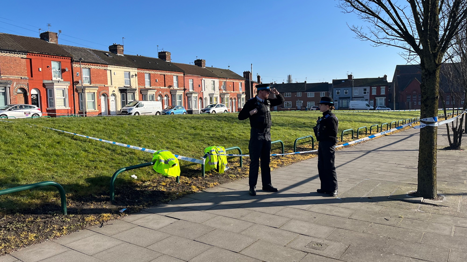 Two police officers stand by a cordon in front of a small grassed area near terraced houses