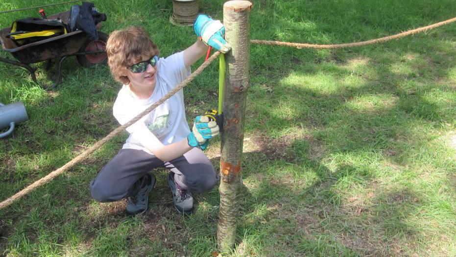 A boy in sunglasses, a white T-shirt and grey tracksuit bottoms, measures part of a tree trunk fence post as it's being installed on a grass field.