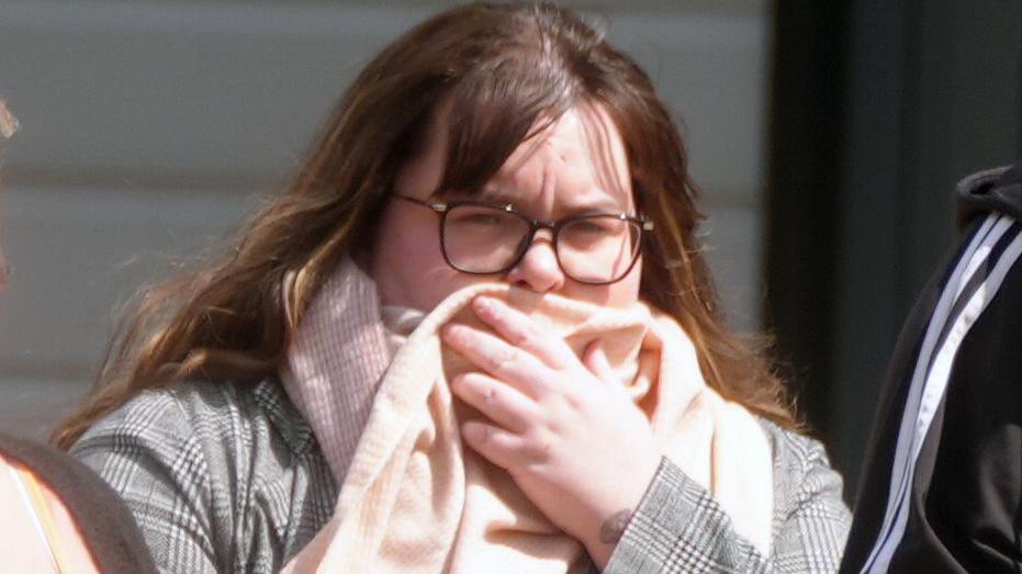 Kelsey Calvert with pink scarf covering him mouth. She has long, brown hair and is wear black, large glasses.