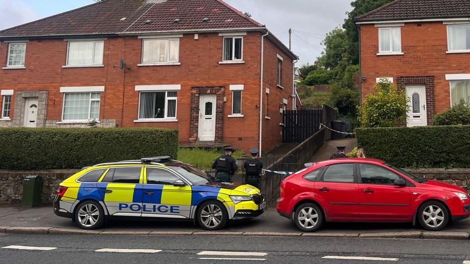 Red brick semi-detached houses. A police cordon is in place at the drive on a house at the right hand side of the picture. In the foreground is a police car and three police officers are standing outside the cordon. There is also a red car in front of the house with the cordon.
