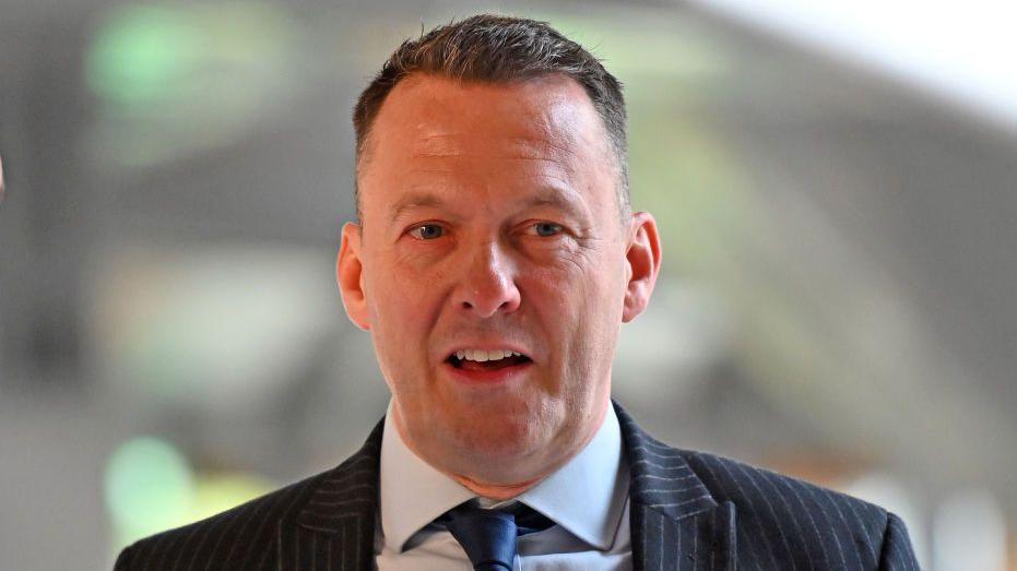 A man with short dark hair, a dark striped suit and blue tie