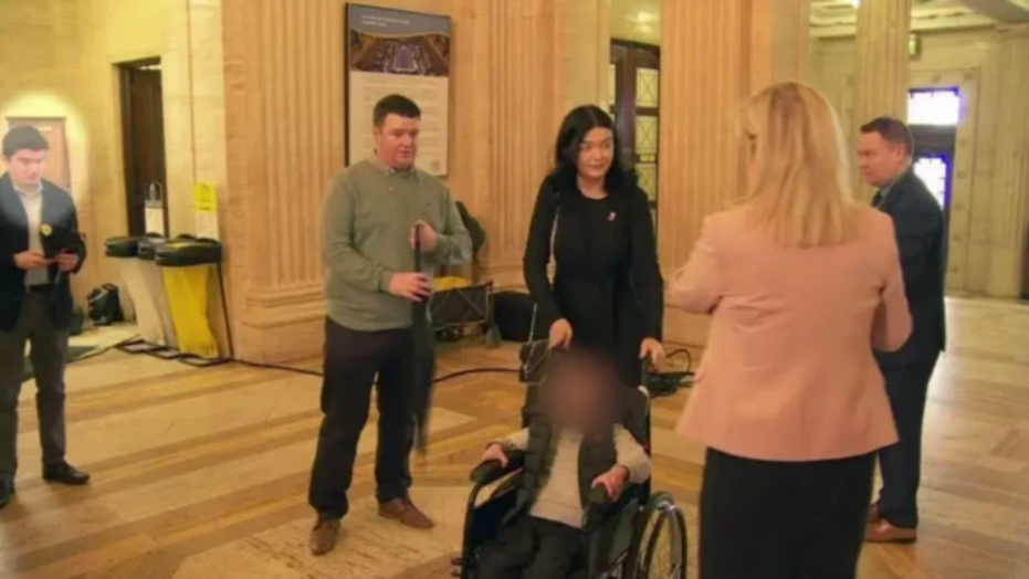 A photo from the event an Stormont showing Michael McMonagle standing to the left of the screen looking down at his phone, he is wearing trousers, shirt and a jacket. Michelle O'Neill stands to the right of the screen with her back to the camera, wearing a pink jacket. In the centre of the image is a child in a wheelchair with their face blurred and their parents standing behind him.