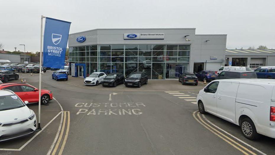 Bristol Street Motors showroom. A load of vehicles are parked beside a large grey building with large windows.