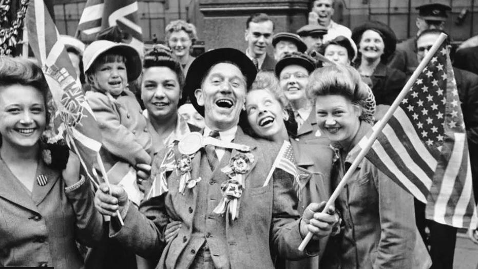 A black and white image of a group of adults and children of varying ages waving flags and smiling at a party to celebrate VE Day in 1945 