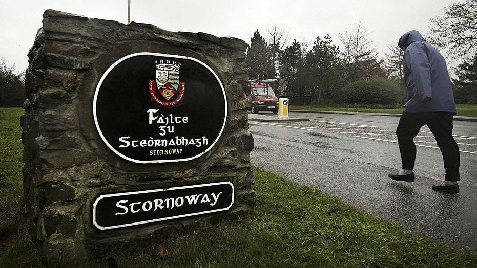 A woman struggles to walk up a pavement in wind. She passes a sign that reads "Stornoway" in Gaelic and English.