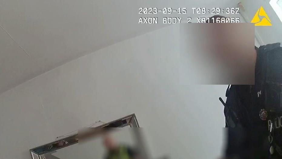 A still images has been taken from the police body-worn camera footage. It shows a police officer standing in her living room. Parts of the image have been blurred to conceal the officer's and woman's identities.
