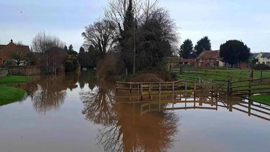 Flooding in Herefordshire - a street and field are covered in water, which is lapping up the grass banks, with homes in distance