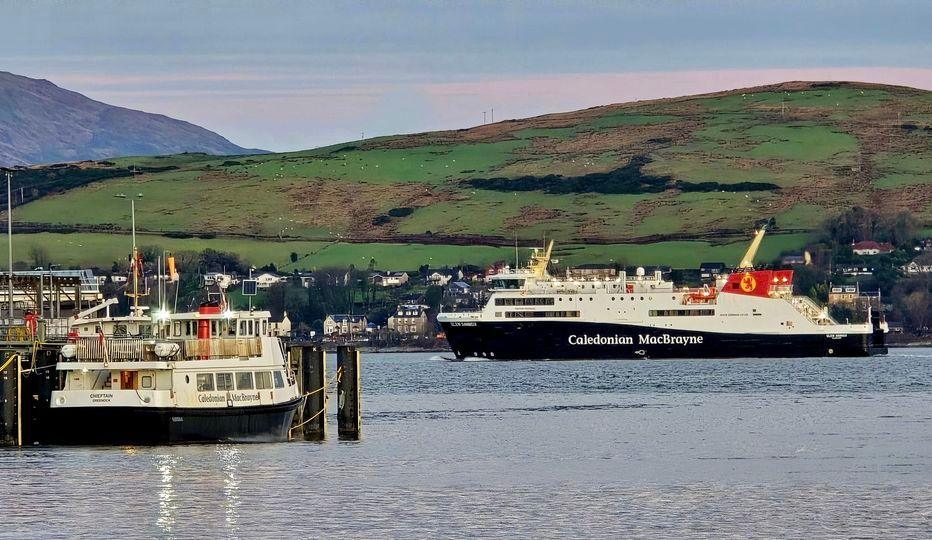 A large black and white ship with red funnels, sailing from right to left on the Clyde, with a much smaller CalMac ship on the left foreground of the picture. 