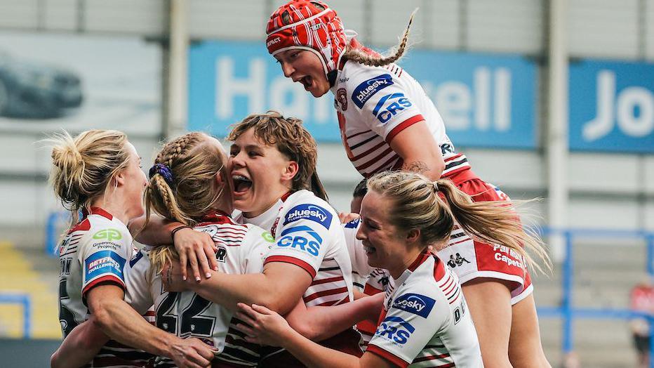 Wigan Warriors women players, in white kit with red trim, surround try-scorer Ruby Hunter, with one jumping on the shoulders of a team-mate