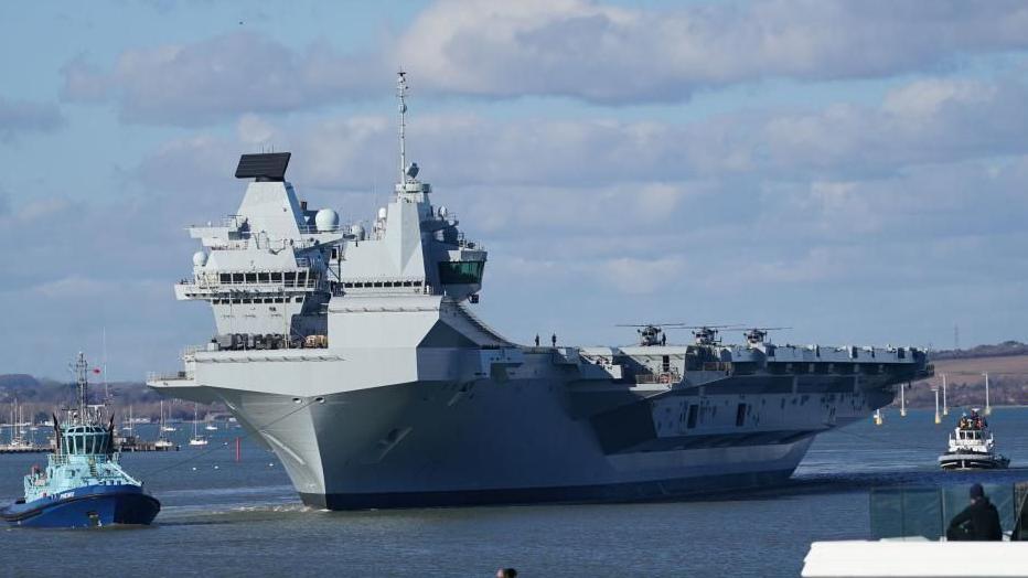 HMS Prince of Wales, an enormous, grey vessel with three helicopters visible on its flight deck, is towed along the Mersey Estuary by a blue tug boat