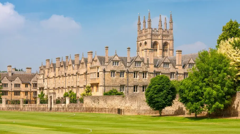 Merton College can be seen on a sunny day, with sports pitches to the front and trees to the right.