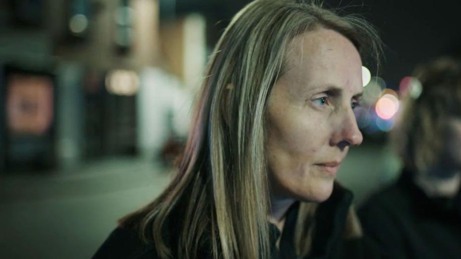 Crime scene coordinator, Jo Ward, is standing in a street. We see only the right side of her face. She has shoulder length blonde hair and is wearing a black top. She is listening attentively to someone who is not in the shot. In the background, you can see properties. it is evening time and the night sky behind Jo is dark.