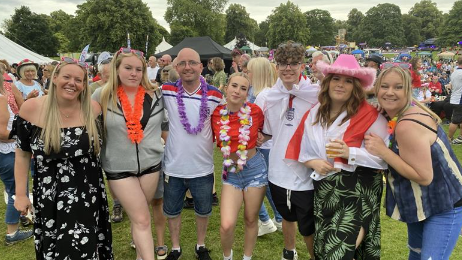 A group of seven fans stand together and smile for the camera, they are at a busy festival with lots of tents and people visible behind them