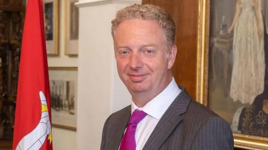 Alex Allinson smiling at the camera. He has fair hair and is wearing a grey suit, white shirt and pink tie, and is standing next to a red Manx flag.