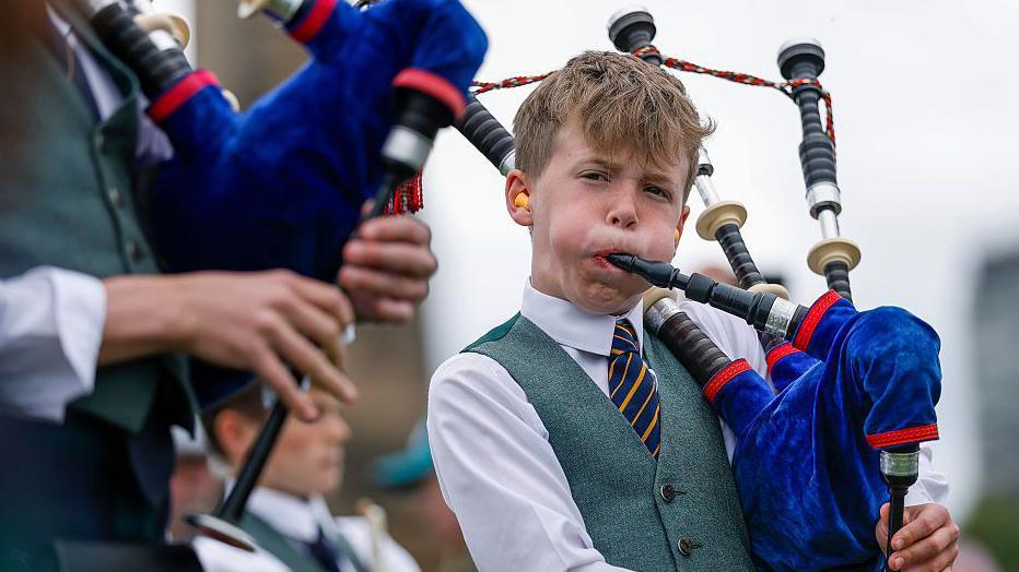 A young male piper playing the bagpipes. His cheeks are puffed out as he plays. He is wearing a traditional piper outfit 