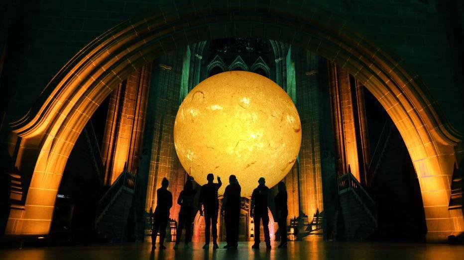 An artist's impression of the sun sculpture called Helios as it will look in Liverpool Cathedral with people standing in front of it pointing to it