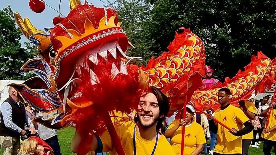 A man, Luke McFarlane at Mela festival pictured with a Chinese dragon. 

He is smiling, has long brunette hair and is wearing a yellow t-shirt. 

Behind him are other performers and trees.