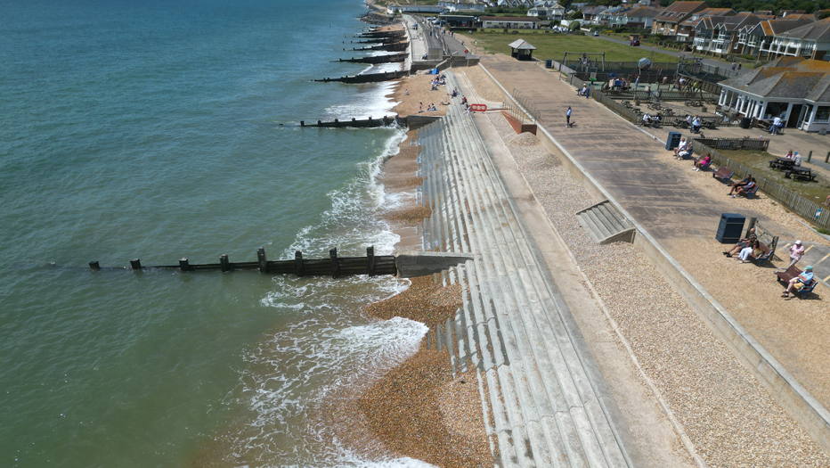 An aerial shot of a shingle beach with wooden groynes placed along it. Behind the beach is a concrete promenade and a square of grassland. Some houses lie beyond it.