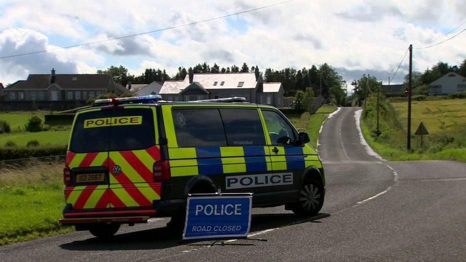 A police van parked on a rural road leading to the crash scene in July 2020. A "police road closed" sign is placed beside the van.  There are a number of houses along the road. 