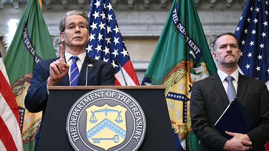 Treasury Secretary Scott Bessent and Trade Representative Jamieson Greer take questions from reporters in Washington DC. The pair are speaking behind a lectern with a prominent US Department of the Treasury plaque displayed.