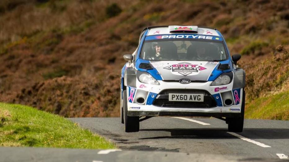 A blue and white rally car races on a Manx road. You can also see the driver and the passenger with helmets through the windscreen.