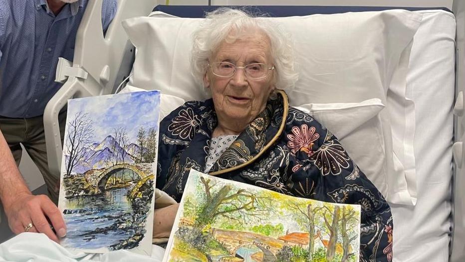 Jean Scarrott, 96, in a hospital bed holding up two landscape paintings. 