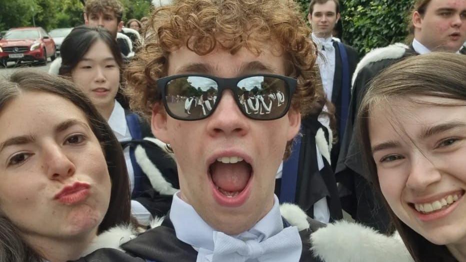 Macsen Brown yn gwisgo gwisg graddio Caergrawnt tra'n cerdded i'r seremoni gyda'i ffrindiau. Mae ganddo wallt coch cyrliog ac mae'n gwisgo sbectol haul du.