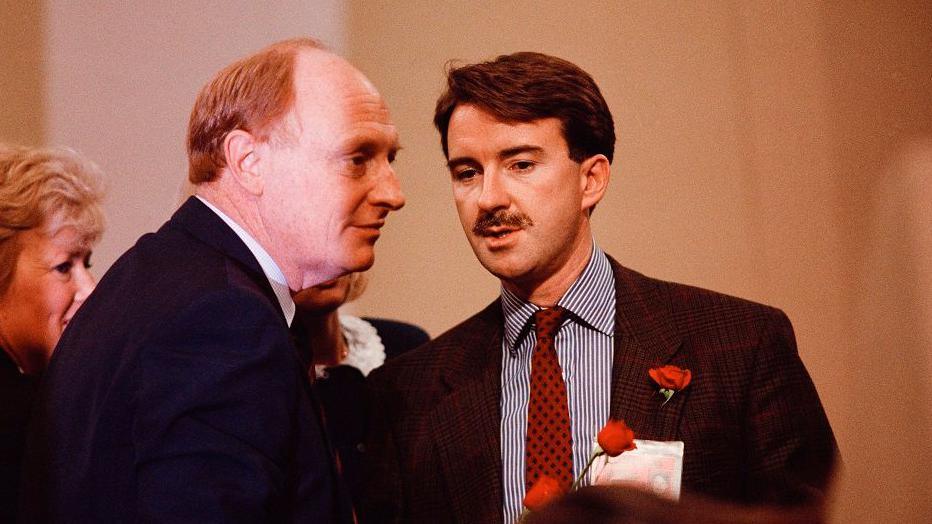 Then Labour leader Neil Kinnock confers with a moustachioed Peter Mandelson in 1989. Both men are wearing dark suits