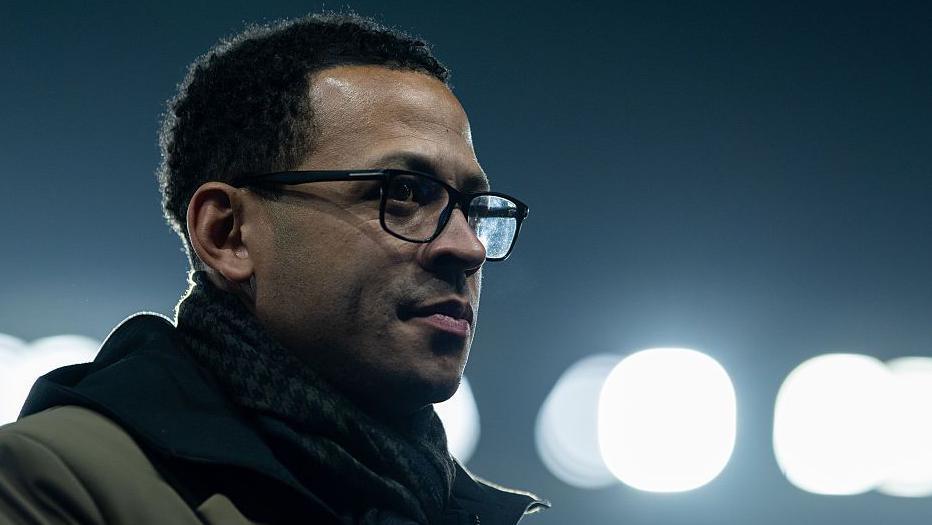 Liam Rosenior in black-rimmed glasses, dark scarf and coat, with lights illuminating a dark background