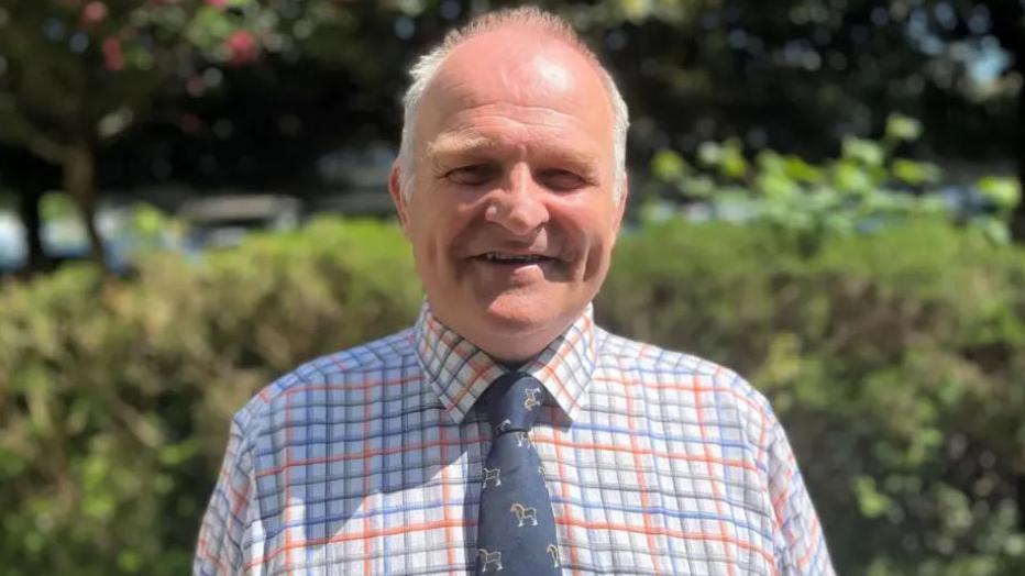 Huw Murphy pictured outside in sunshine wearing white and orange and green checked shirt and navy tie with horses on it. He has short grey hair and is smiling