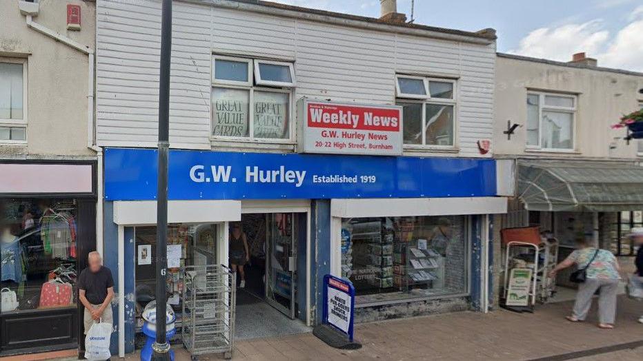 The shopfront on an overcast day. The sign is on blue and white and a few people are walking past or standing nearby.